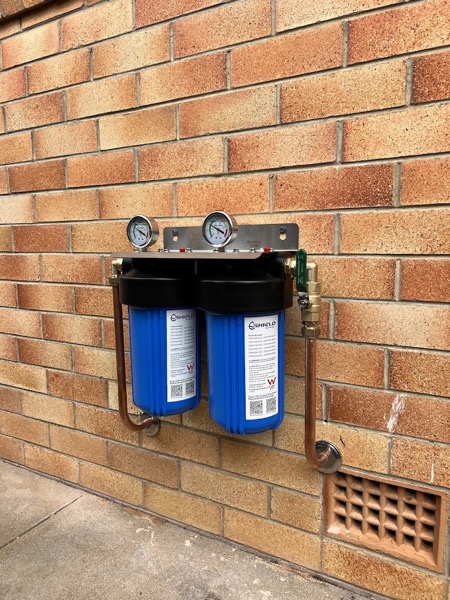A twin cartridge whole-house water filter mounted on a brick wall with pressure gauges