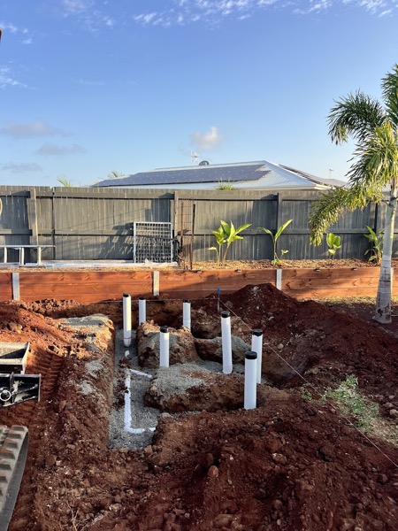 Drainage risers standing in a trench of red soil on a new build, with palms behind