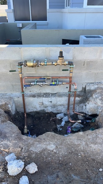 A copper water meter and mains assembly rebuilt in an excavated pit beside a house